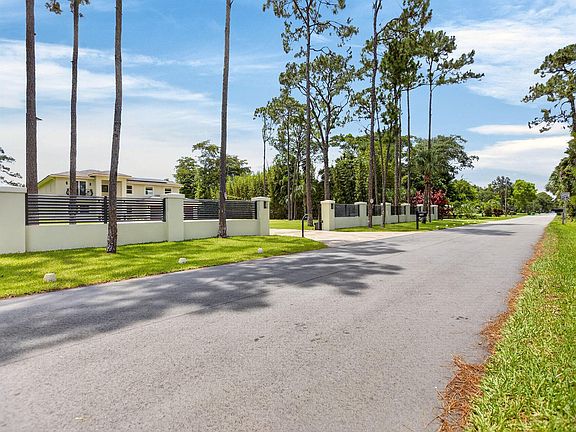 Streetside view of Pine Tree Estates: a wide quiet lane lined with tall pines and slat fencing along the property edge