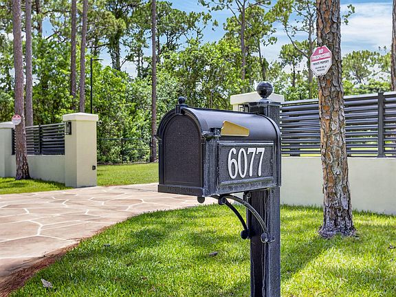 Close detail of the property's gated entry, the address number 6077 visible on the cast mailbox, framed by pine trunks